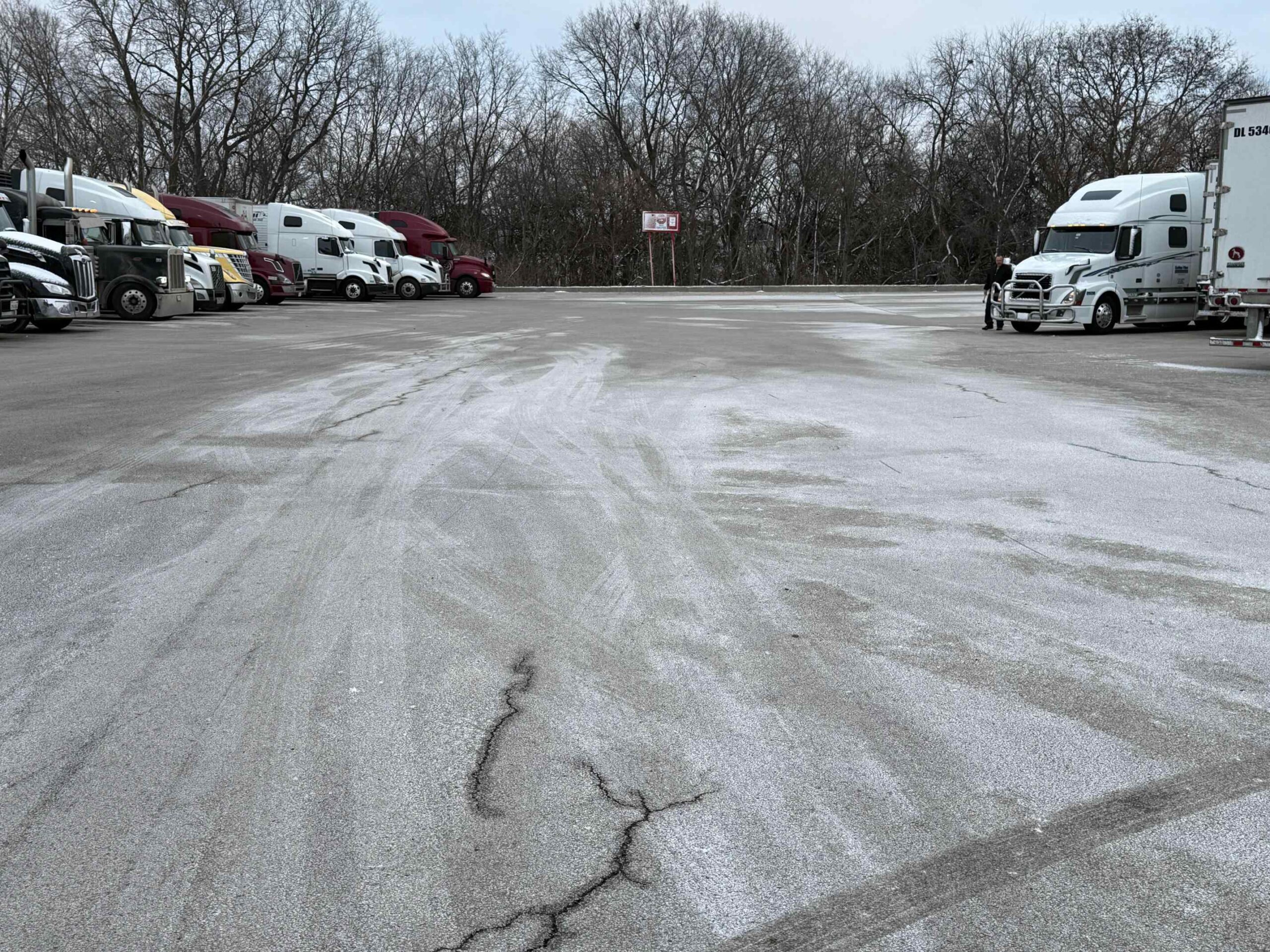 A large commercial asphalt parking lot with parked semi-trucks, showing cleared and salted ground during winter.